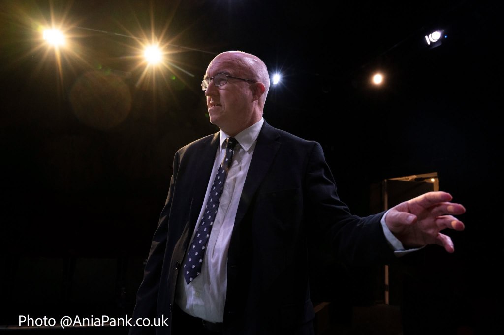 A man in a suit stands on stage with dramatic lighting, gesturing as he speaks.