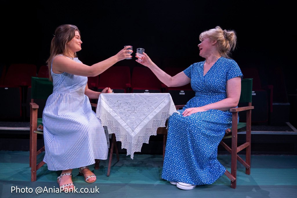 Two women sitting at a small table with a lace tablecloth, raising glasses in a toast, while dressed in summer dresses.